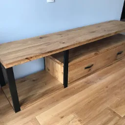 Drewniana szafka RTV z szufladą i czarnymi metalowymi nogami, stojąca na drewnianej podłodze przy jasnoniebieskiej ścianie.