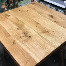 Blat kwadratowego stołu z jasnego drewna dębowego z widocznymi słojami i sękami, sfotografowany pod kątem, z puszkami z farbą w tle.