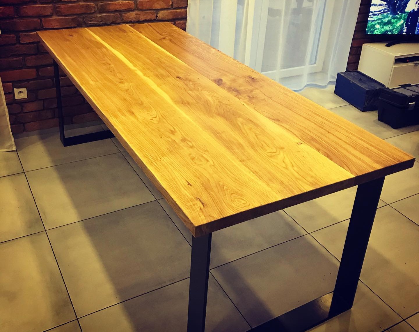 Stół z blatem z jasnego drewna dębowego i czarnymi, metalowymi nogami w nowoczesnym wnętrzu z ceglaną ścianą w tle.