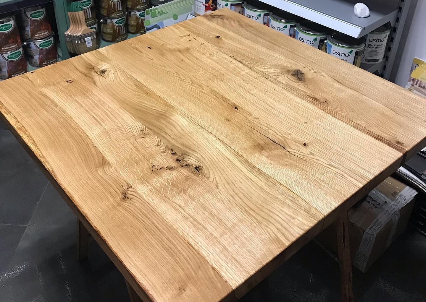 Blat kwadratowego stołu z jasnego drewna dębowego z widocznymi słojami i sękami, sfotografowany pod kątem, z puszkami z farbą w tle.