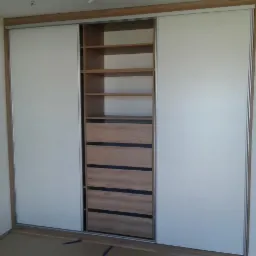 Wnętrze pokoju z szafą przesuwną z białymi drzwiami i drewnianymi półkami oraz szufladami z czarnymi akcentami, widoczne gniazdko elektryczne na ścianie.