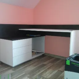 Biało-czarny mebel z szufladami i p&oacute;łką, prawdopodobnie biurko, z narzędziem Festool w rogu pokoju z r&oacute;żowymi ścianami, na drewnianej podłodze.