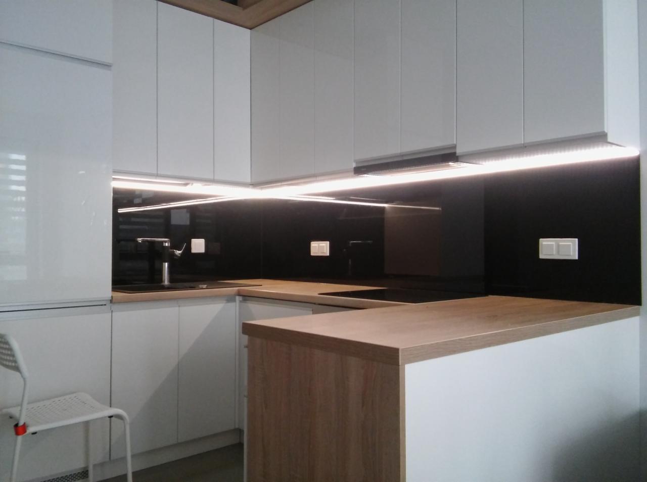 Nowoczesna kuchnia z białymi frontami szafek, czarnym panelem szklanym nad blatem i oświetleniem LED podszafkowym, uzupełniona blatem w kolorze jasnego drewna.