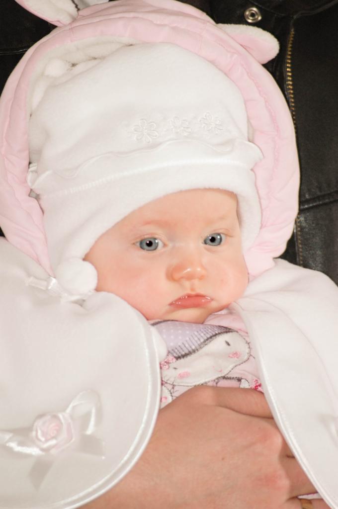 Niemowlę z niebieskimi oczami, ubrane w biały kombinezon z kapturem w kolorze pudrowego różu i aplikacjami kwiatowymi, trzymane na rękach.