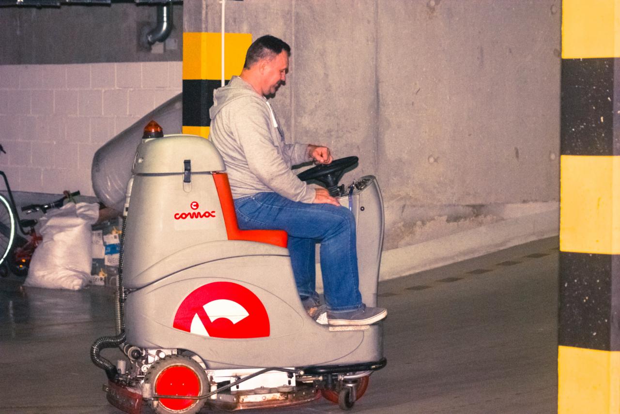 Mężczyzna w szarej bluzie i jeansach obsługuje maszynę czyszczącą podłogi marki Comac w garażu podziemnym.
