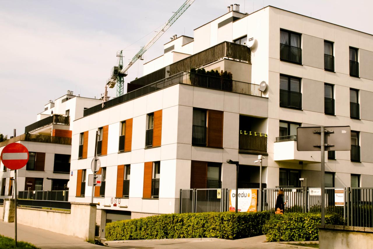 Nowoczesny budynek mieszkalny z balkonami i drewnianymi okiennicami, w tle dźwig budowlany i znak zakazu wjazdu.