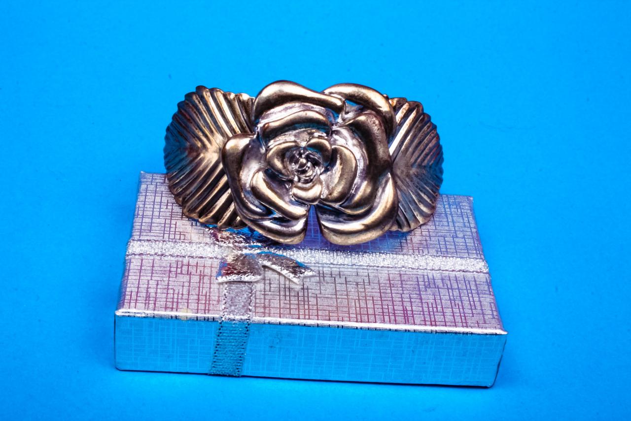 Metalowa broszka w kształcie róży na srebrnym pudełku z kokardką na niebieskim tle.