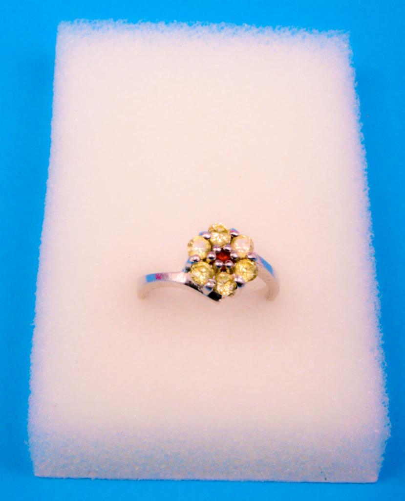 Srebrny pierścionek z żółtymi i czerwonymi kamieniami ułożonymi w kształt kwiatu, prezentowany na białej gąbce na tle niebieskim.