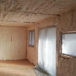 Wnętrze pomieszczenia po natrysku izolacji, ściany i sufit pokryte grubą warstwą pianki, okna zabezpieczone folią, widoczny karton i fragment podłogi.