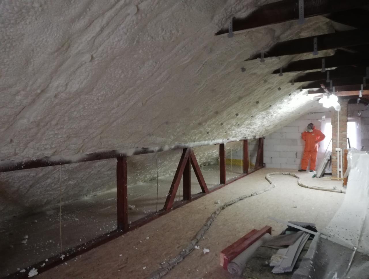 Poddasze po natrysku izolacji, widoczna gruba warstwa piany na skośnym dachu i pracownik w kombinezonie ochronnym w tle.