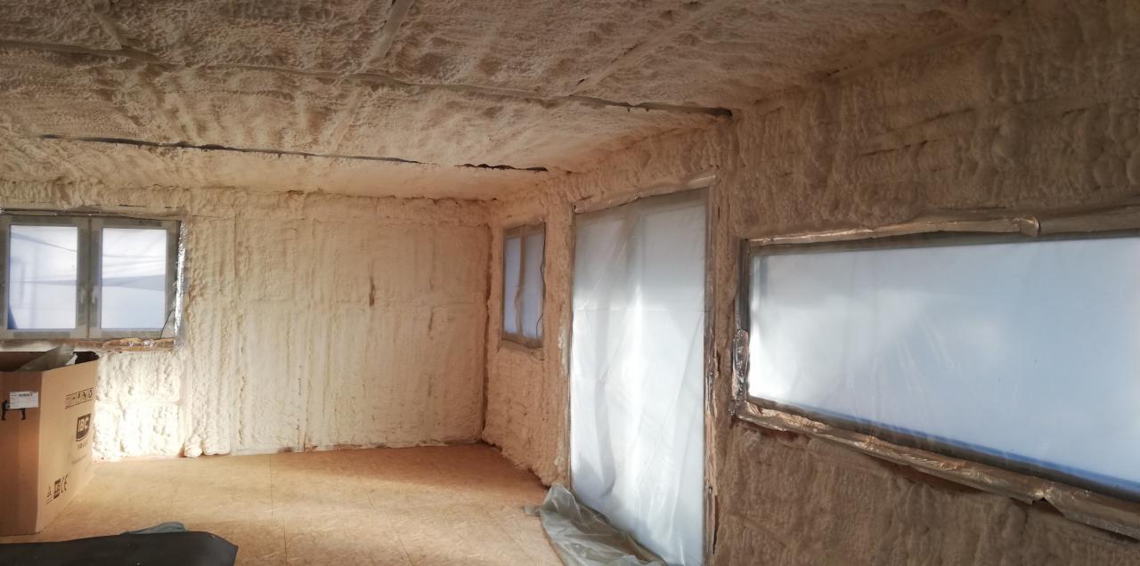 Wnętrze pomieszczenia po natrysku izolacji, ściany i sufit pokryte grubą warstwą pianki, okna zabezpieczone folią, widoczny karton i fragment podłogi.