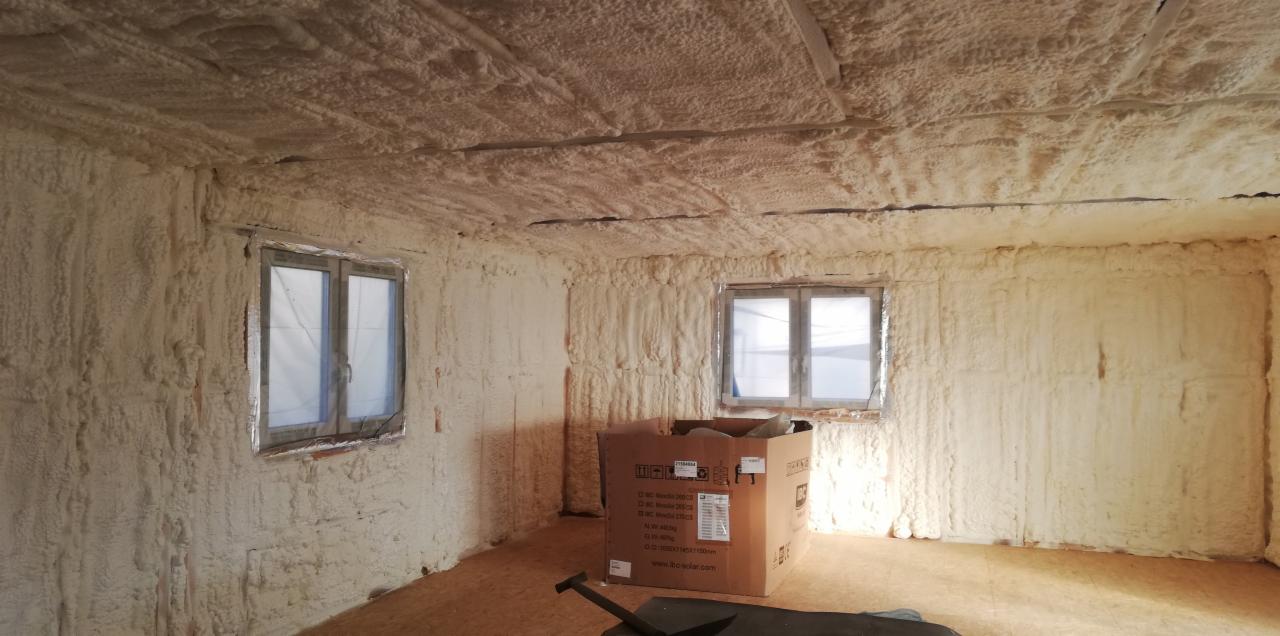 Wnętrze pokoju po ociepleniu pianką PUR, widoczne okna zabezpieczone folią, karton na podłodze i narzędzie do docinania pianki.
