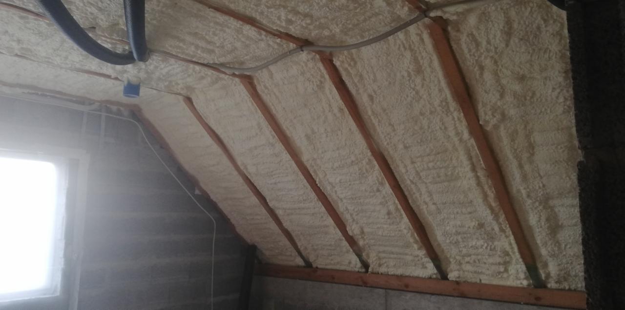 Poddasze ocieplone pianą, widoczne drewniane krokwie i instalacje elektryczne, szara ściana z oknem w tle.