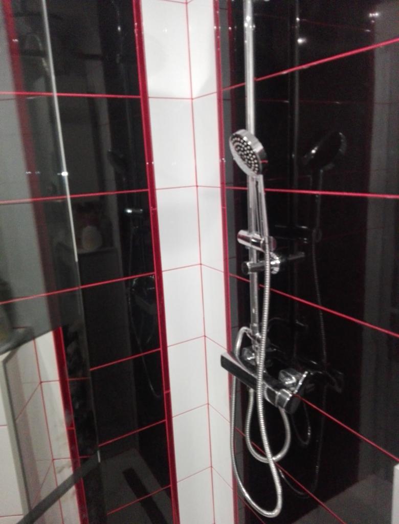 Bateria prysznicowa z chromowaną słuchawką na tle ściany wyłożonej czarno-białymi płytkami z czerwoną fugą.