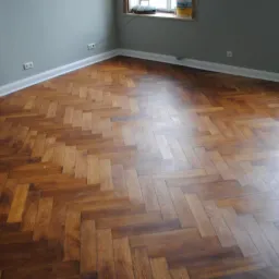 Parkiet ułożony w jodełkę po cyklinowaniu, widoczne ślady po procesie oraz puszka z lakierem na parapecie okna.