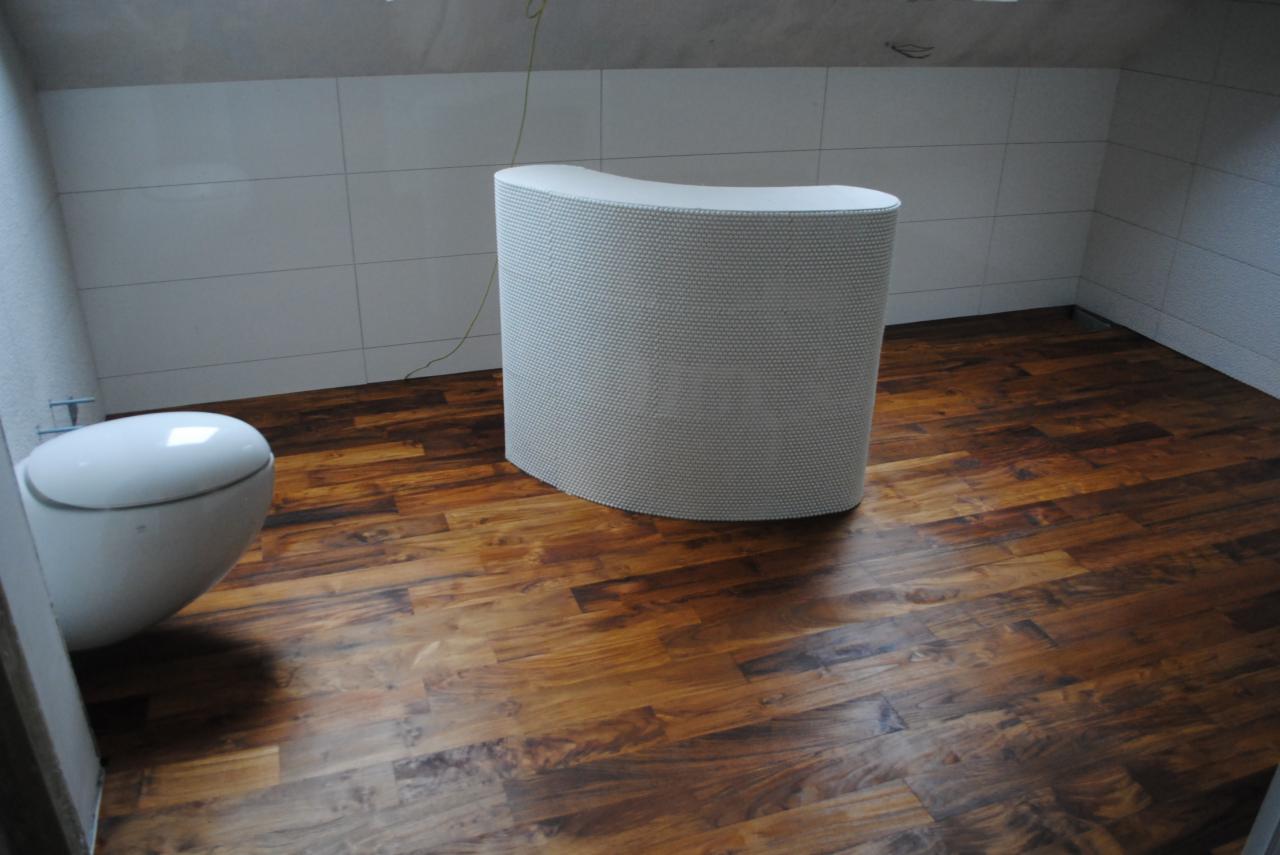 Nowa, ciemna, błyszcząca drewniana podłoga w łazience z białą toaletą i obudową rur.