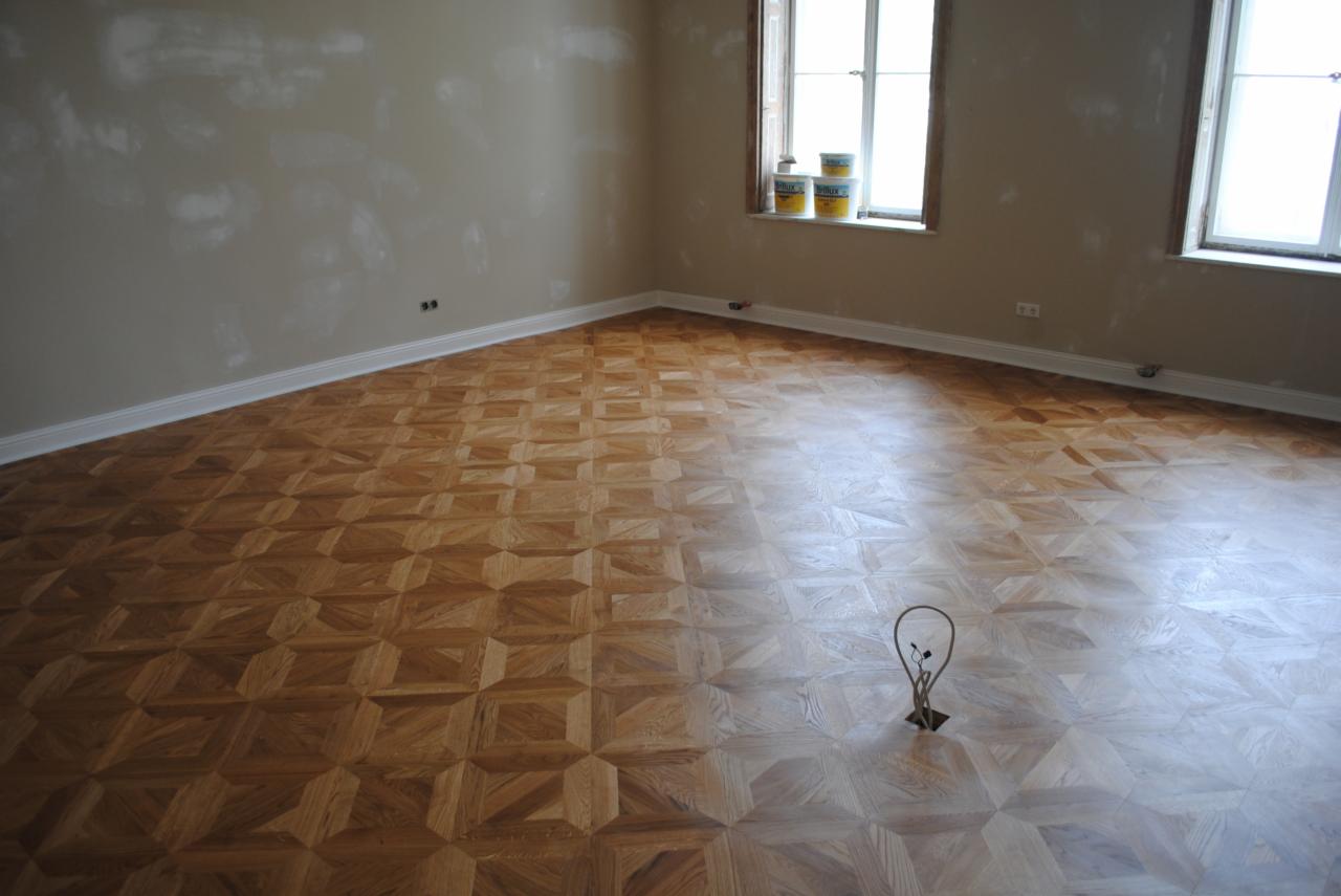 Parkiet ułożony we wzór szachownicy w jasnym pokoju, ściany przygotowane do malowania, puszki z farbą na parapecie okna.