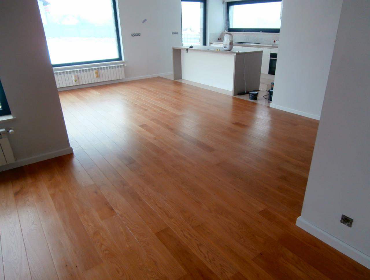 Salon z ułożoną, lakierowaną podłogą z desek o ciepłym, miodowym odcieniu, widok na kuchnię z białymi meblami i okno z widokiem na zewnątrz.