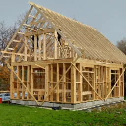 Surowy szkielet drewniany domu w trakcie budowy, widoczne elementy konstrukcyjne dachu i ścian, pracownicy na g&oacute;rze, fundament betonowy, zielona trawa wok&oacute;ł.