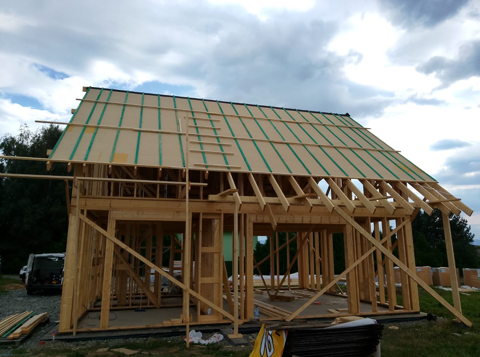 Wznoszenie szkieletu drewnianego domu z widoczną konstrukcją dachu, zielonymi elementami izolacyjnymi i stertą materiałów budowlanych na trawie.