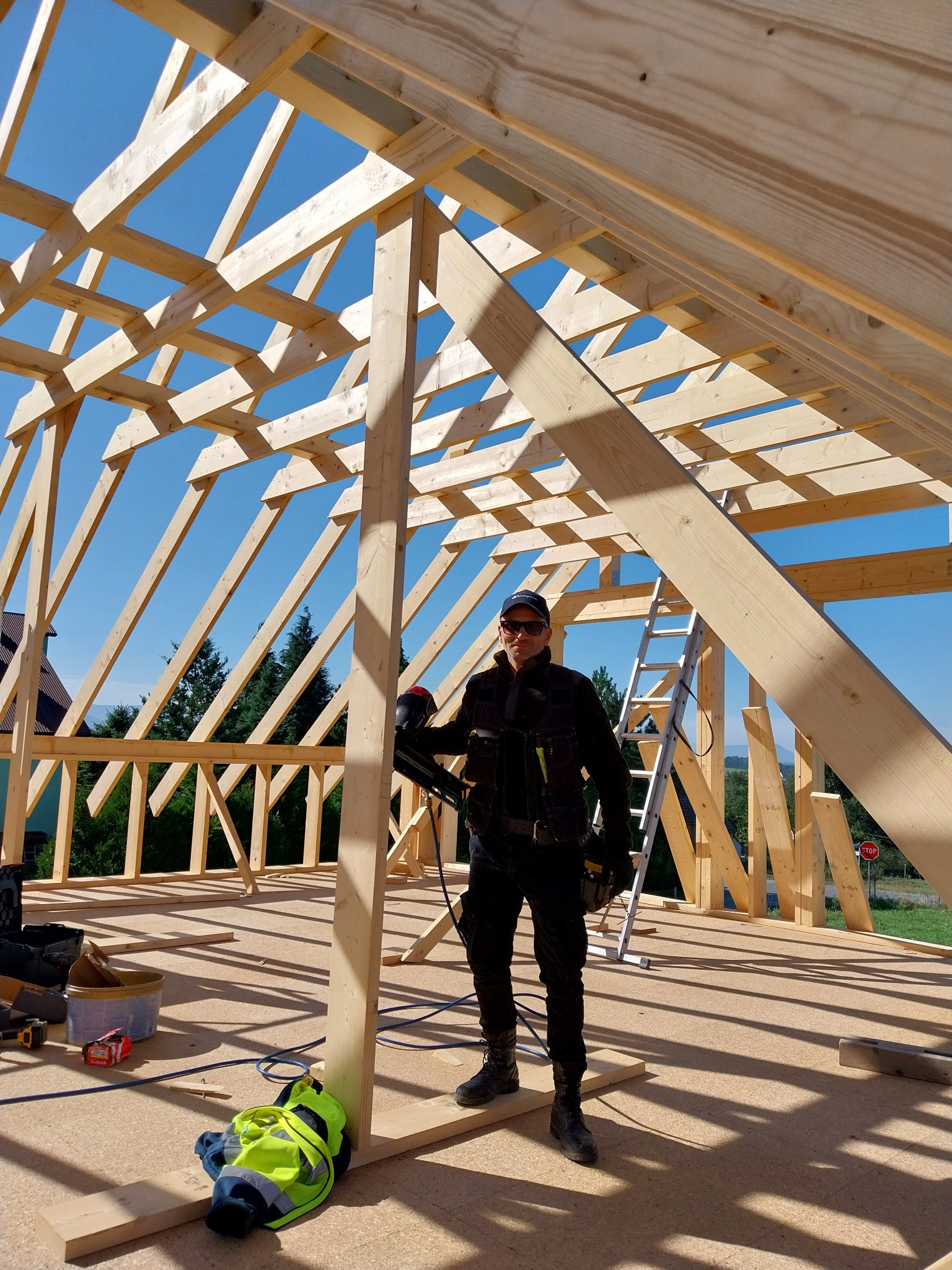 Konstrukcja drewniana domu w trakcie budowy, widoczny dekarz z narzędziem do montażu więźby dachowej na tle błękitnego nieba.