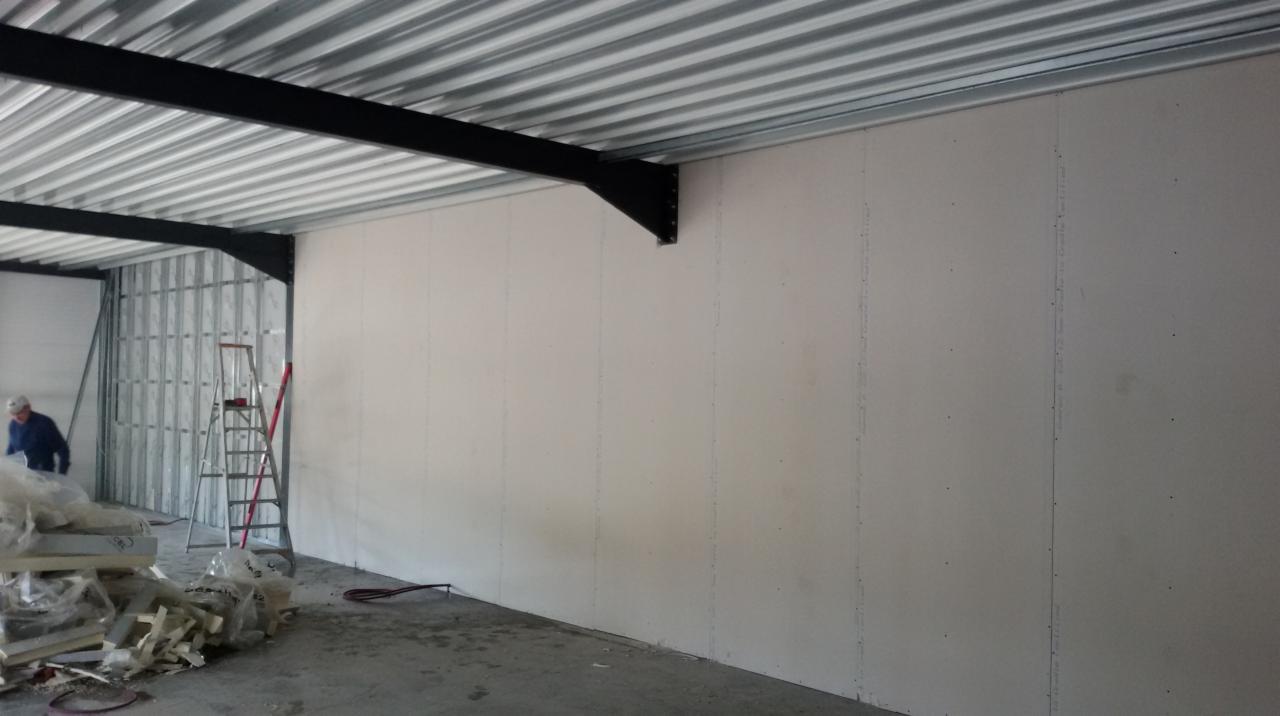 Montaż płyt gipsowo-kartonowych na ścianie w nowym biurze, widoczna konstrukcja metalowa sufitu i ściany, drabina i materiały budowlane w tle, pracownik w oddali.