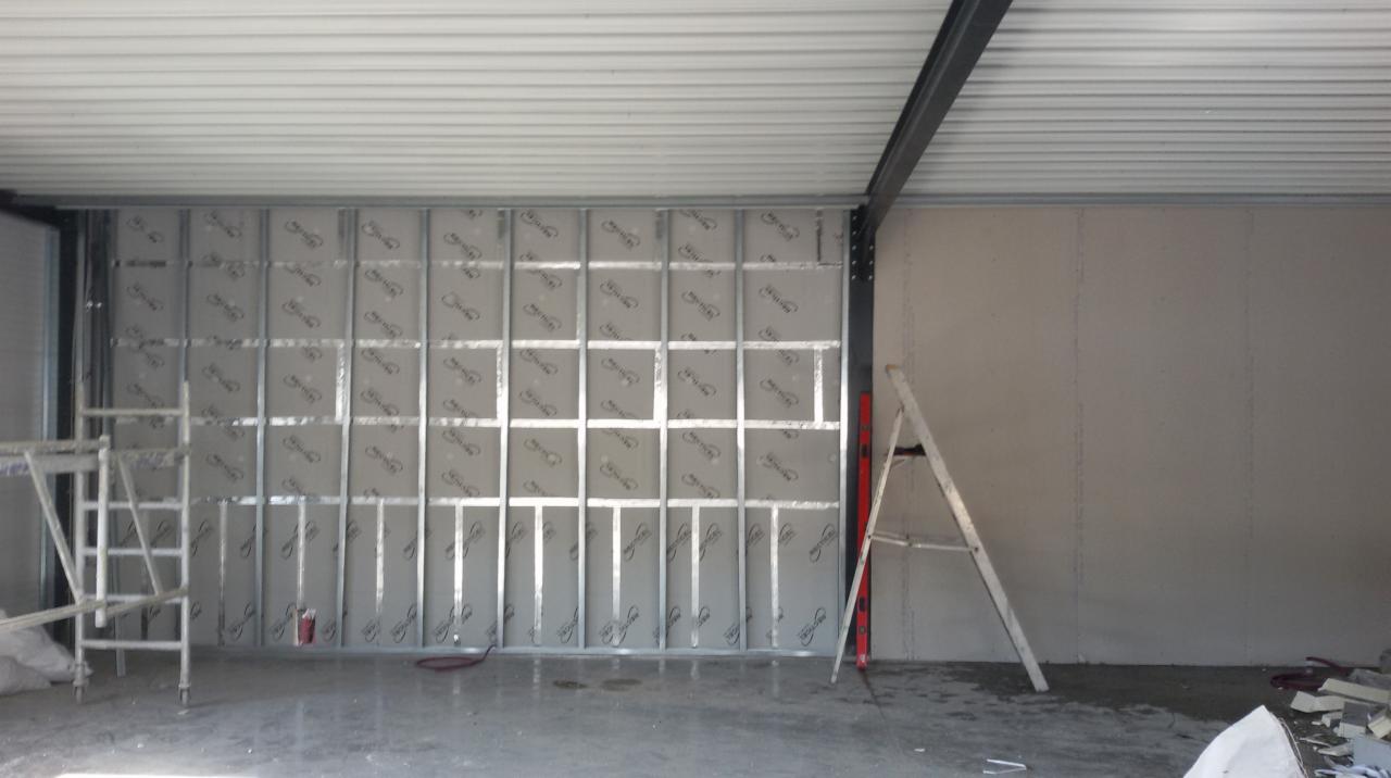 Montaż konstrukcji metalowej pod płyty gipsowe w dużej hali, widoczny ruszt i częściowo zamontowane płyty z nadrukiem producenta, drabina oparta o ścianę.