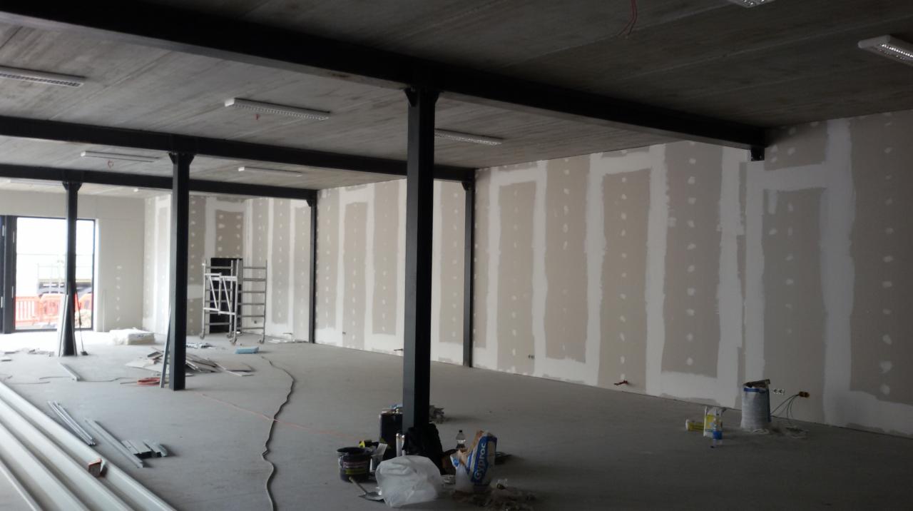 Wnętrze biurowe w trakcie wykańczania, widoczne płyty kartonowo-gipsowe na ścianach, metalowe słupy konstrukcyjne, surowy betonowy sufit z lampami i akcesoria budowlane na podłodze.