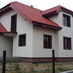 Dwukondygnacyjny dom z beżową elewacją, brązowymi ramami okiennymi i czerwoną dachówką, widziany z zewnątrz w pochmurny dzień.