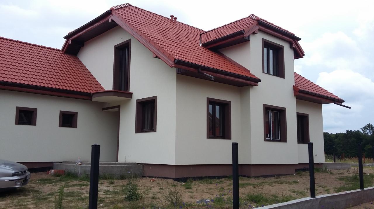 Dwukondygnacyjny dom z beżową elewacją, brązowymi ramami okiennymi i czerwoną dachówką, widziany z zewnątrz w pochmurny dzień.