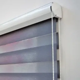 Szaro-różowa roleta z poziomymi pasami, delikatnie przepuszczająca światło, zamontowana w białej kasecie z widocznym mechanizmem łańcuszkowym.