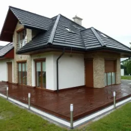 Nowoczesny dom jednorodzinny z ciemną dachówką i elewacją z elementami kamienia, z drewnianym tarasem i brązowymi roletami zewnętrznymi na oknach, widok z zewnątrz.