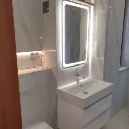 Nowoczesna łazienka z marmurowymi płytkami, podświetlanym lustrem nad białą szafką z umywalką i wiszącą toaletą Geberit.