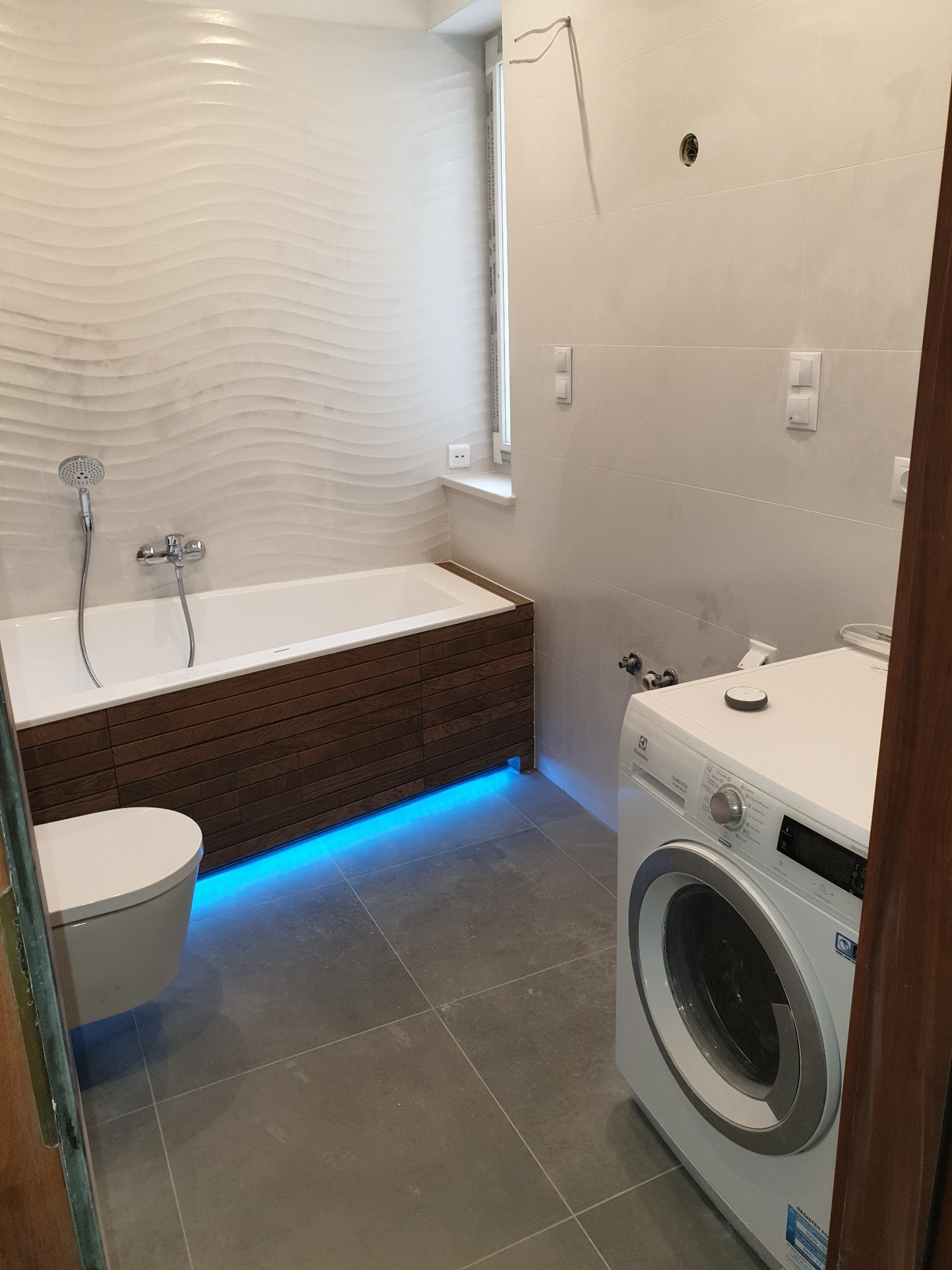 Nowoczesna łazienka po remoncie z wanną obudowaną drewnem i podświetleniem LED, wiszącą toaletą i pralką Electrolux.