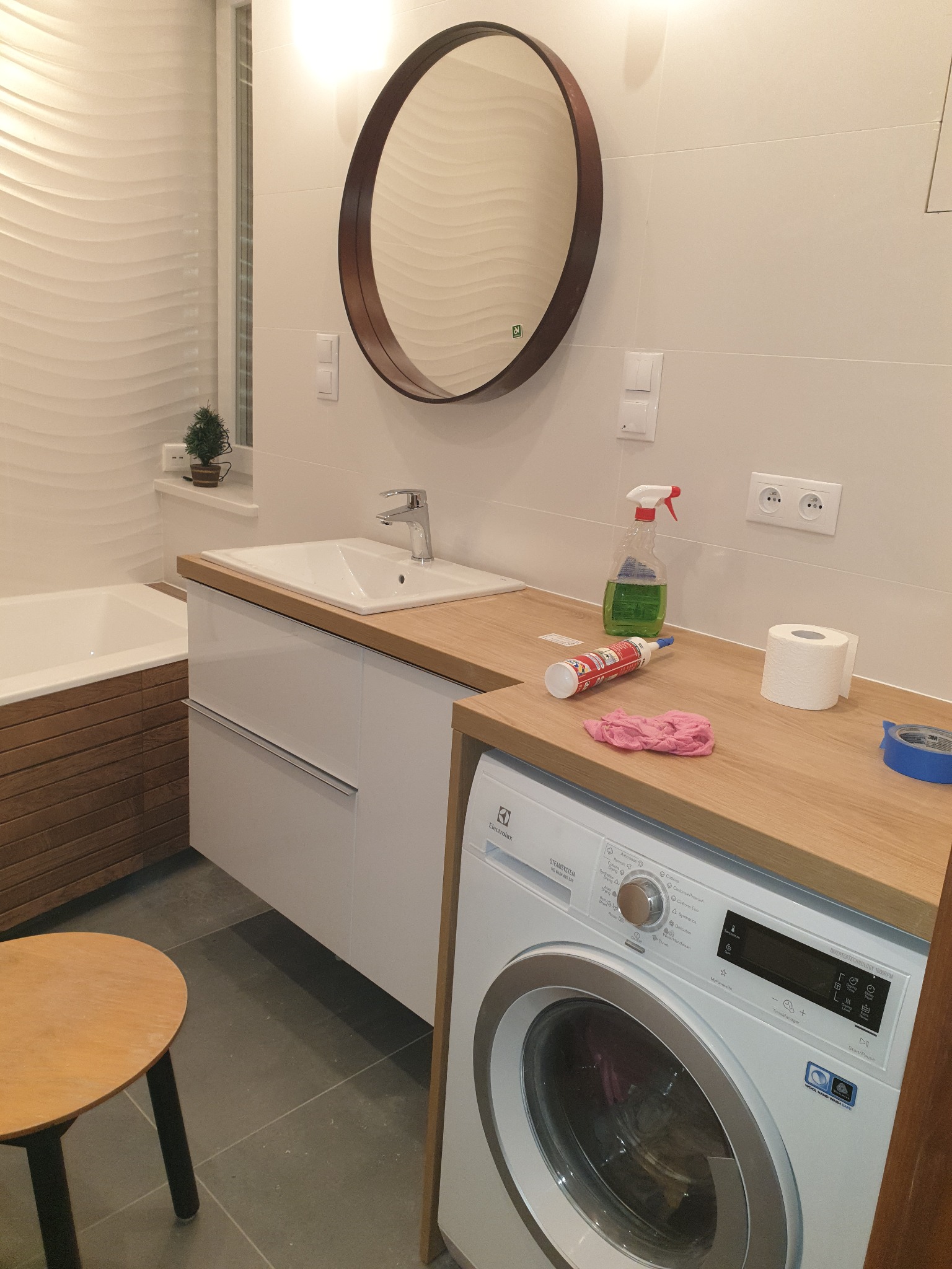 Nowoczesna łazienka po remoncie z okrągłym lustrem, umywalką w szafce, pralką Electrolux i akcesoriami na blacie.