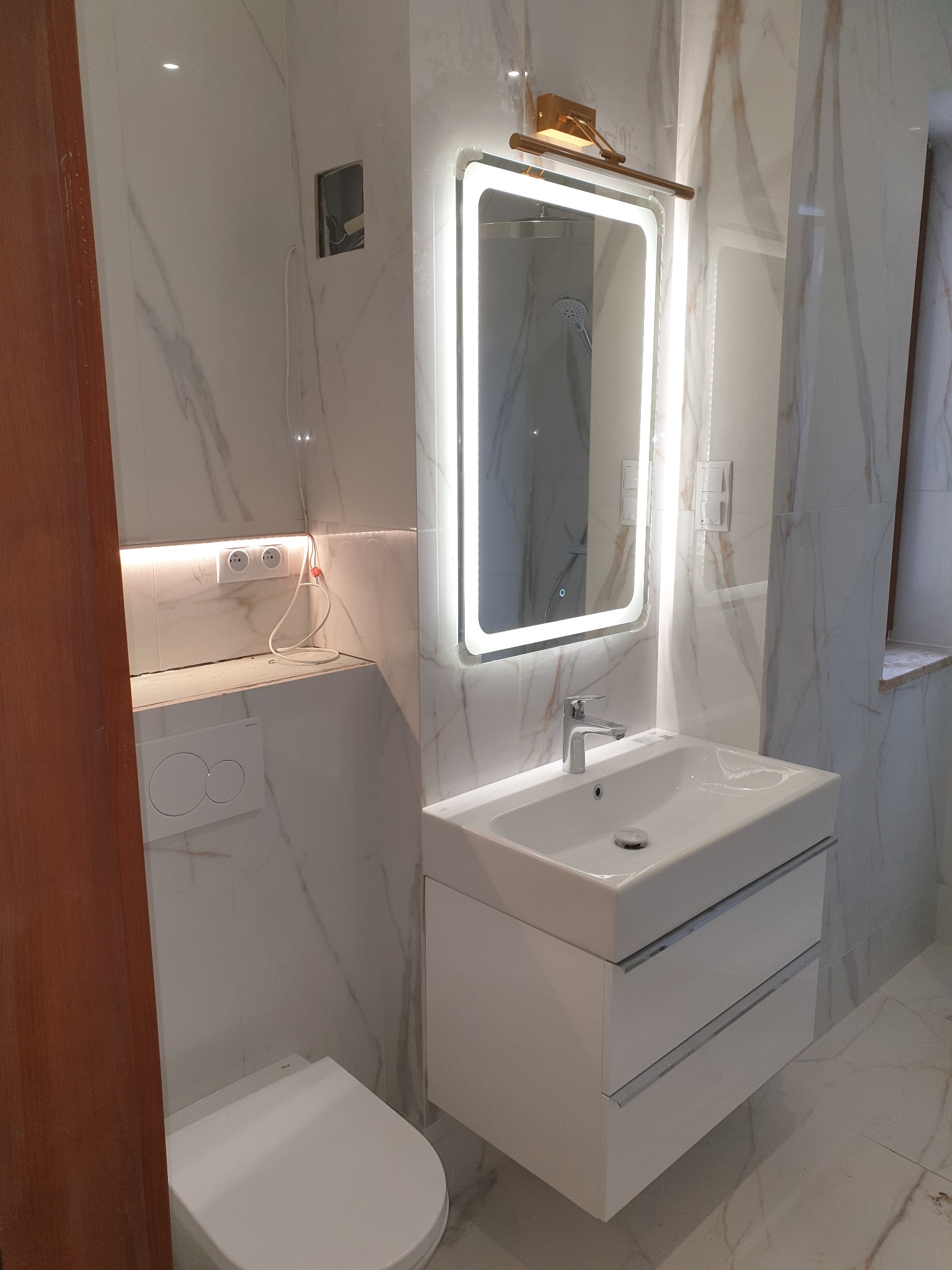 Nowoczesna łazienka z marmurowymi płytkami, podświetlanym lustrem nad białą szafką z umywalką i wiszącą toaletą Geberit.