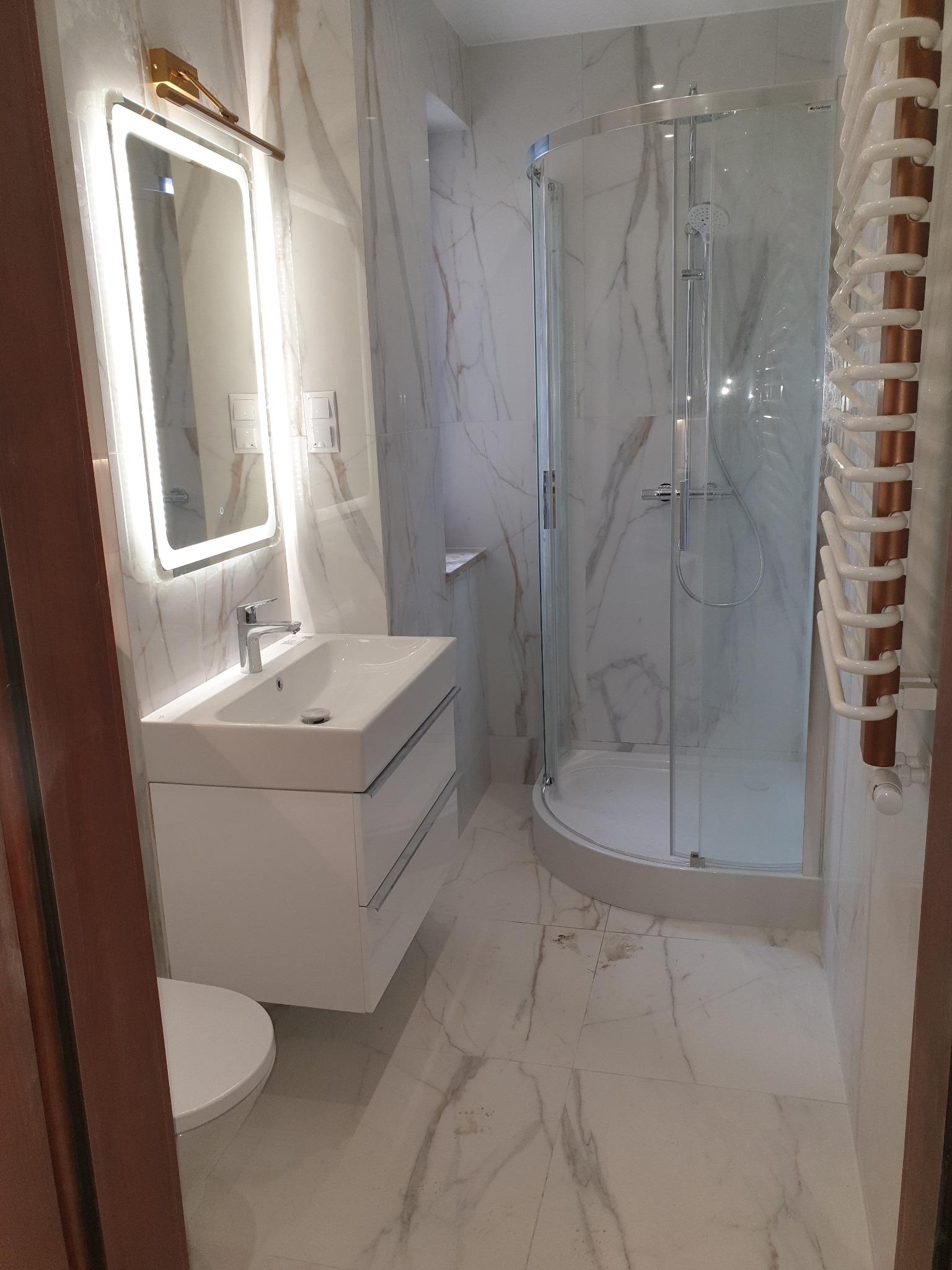 Wykończona łazienka z marmurowymi płytkami, kabiną prysznicową z zaokrąglonymi drzwiami, umywalką z szafką, podświetlanym lustrem i grzejnikiem drabinkowym.