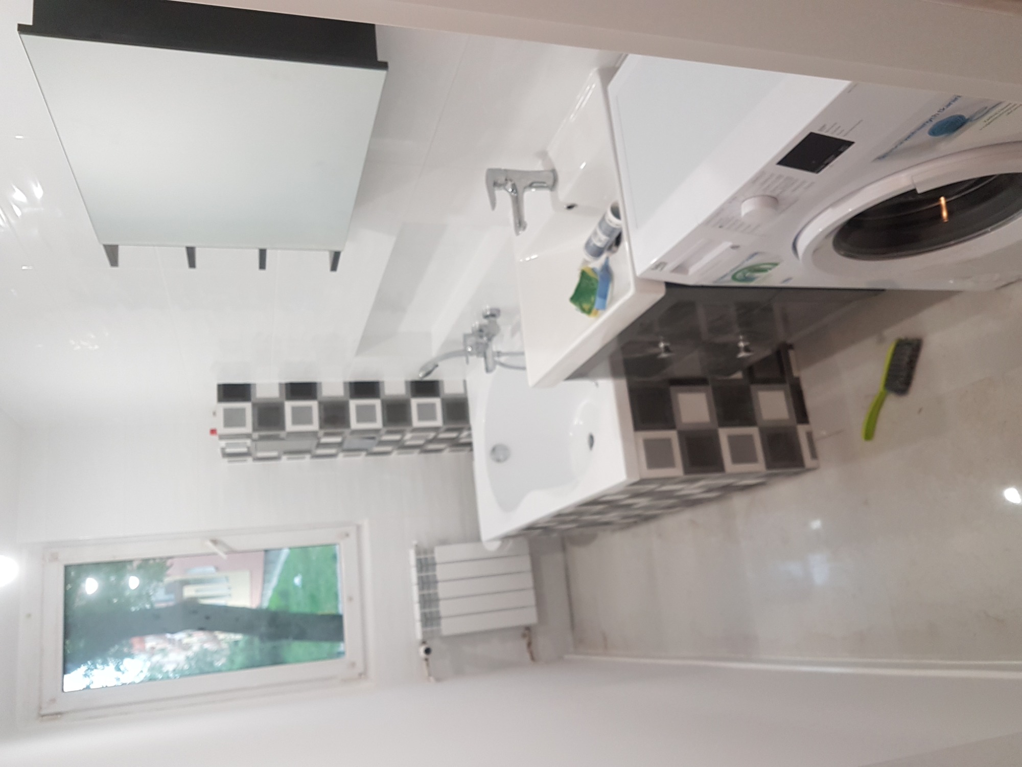 Nowoczesna, biała łazienka z pralką, umywalką, wanną i grzejnikiem, wykończona płytkami w szaro-białą szachownicę.