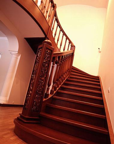 Zakręcone, ciemne, drewniane schody z bogato zdobioną balustradą, widoczne z dołu, z parkietem na podłodze i białymi ścianami w tle.