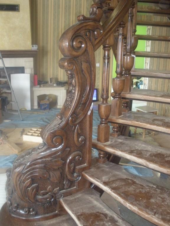 Fragment dębowej balustrady z rzeźbionymi motywami roślinnymi, widoczne niedokończone stopnie i elementy w trakcie montażu na tle pokoju z tapetą w paski.