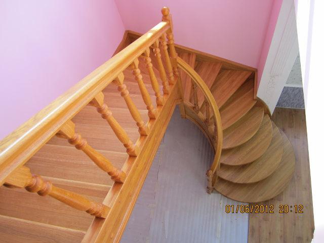 Widok z góry na drewniane schody z zakrętem i balustradą z toczonymi elementami, na tle różowej ściany i fragmentu podłogi.