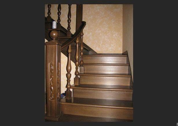 Klasyczne, ciemnobrązowe schody z drewna z ozdobną balustradą i rzeźbionym słupkiem początkowym, częściowo oświetlone, prowadzące w górę.