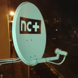 Biała antena satelitarna z logo nc+ zamontowana na balkonie, widok nocny na miasto.