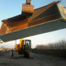 Żółta koparka gąsienicowa z bardzo dużą łyżką, prawdopodobnie transportująca materiał sypki na nierównym terenie, widoczna w oddali roślinność.