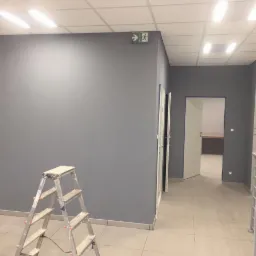 Świeżo pomalowane szare ściany w biurze, widoczna aluminiowa drabina malarska z drobnymi śladami farby, podświetlony znak ewakuacyjny na ścianie, otwarte drzwi do kolejnego pomieszczenia.