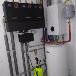 Instalacja grzewcza Bosch z kotłem gazowym, zasobnikiem ciepłej wody i rozdzielaczem, widoczny również opryskiwacz ciśnieniowy w pomieszczeniu technicznym.