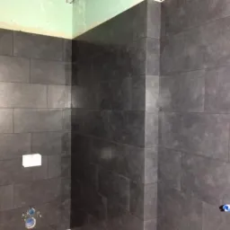 Wykończona ściana łazienki z szarych, prostokątnych płytek ceramicznych, widoczne niezakończone prace wykończeniowe przy suficie i wystające rury instalacji wodnej.