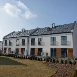 Nowoczesne, dwupiętrowe segmenty mieszkalne z szarym dachem i białymi elewacjami, widoczne okna dachowe oraz balustrady balkonowe z przezroczystego szkła, otoczone zielonym trawnikiem i młodymi...