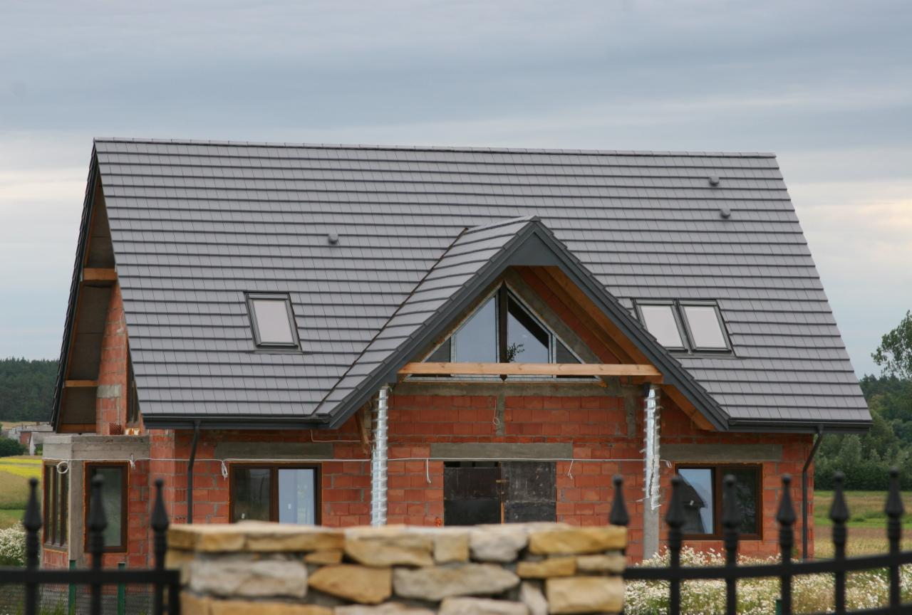 Niedokończony dom z szarą dachówką, widoczne okna dachowe, surowa cegła i elementy konstrukcyjne, zabezpieczone otwory okienne i drzwiowe, fragment ogrodzenia w pierwszym planie.