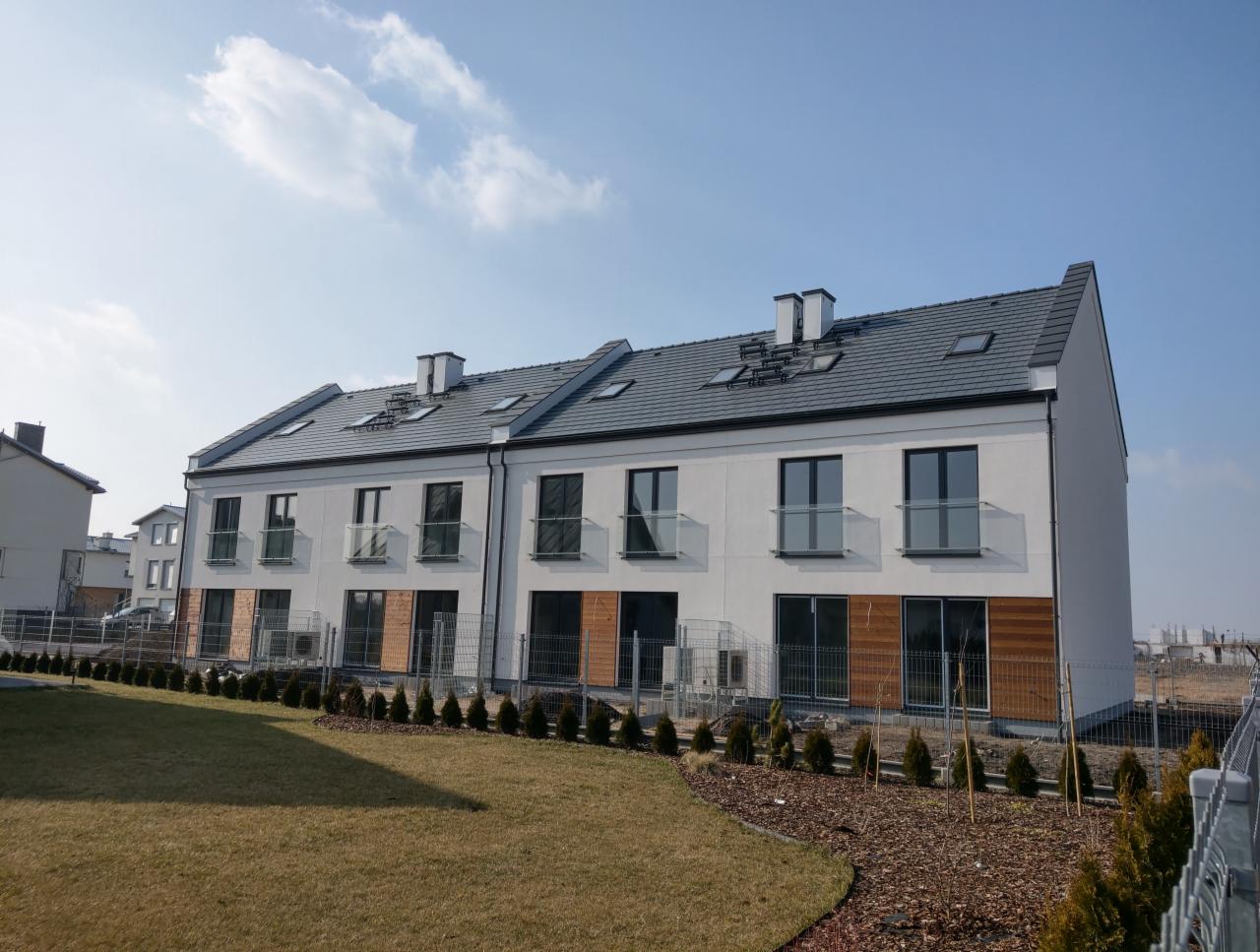 Nowoczesne, dwupiętrowe segmenty mieszkalne z szarym dachem i białymi elewacjami, widoczne okna dachowe oraz balustrady balkonowe z przezroczystego szkła, otoczone zielonym trawnikiem i młodymi...